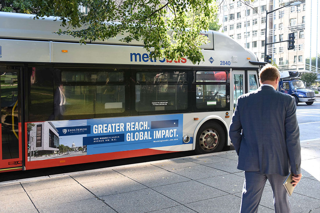 A tall man in a suit approaches a Metro Bus on a sidewalk in Washington, D.C. The advertisement on the side of the bus includes a picture of Johns Hopkins University