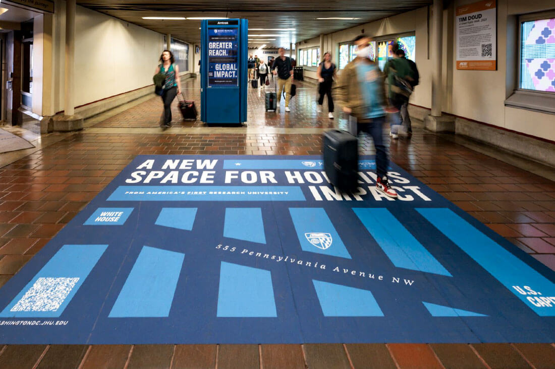 In a long hallway, commuters carrying luggage walk across a giant floor decal advertisement for Johns Hopkins University that includes a line-drawn map of Washington, D.C. The ad reads 'A New Space for Hopkins Impact.' In the background, there's a poster that reads 'Greater Reach. Global Impact.'