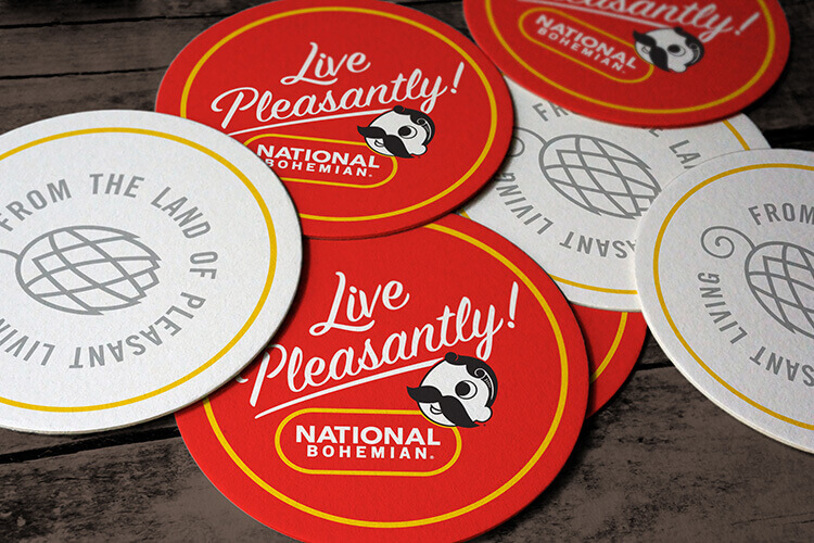 A coaster. One side features the “Live Pleasantly!" slogan and National Bohemian logo and icon, and the other side features an icon of a hop with additional brand messaging.
