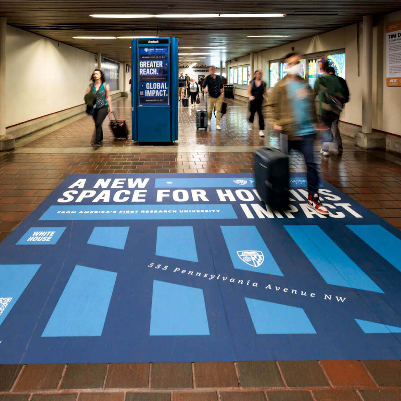 A large-scale decal advertisement on the tiled floor of a D.C. transit hub. A digital advertisement can be seen in the background.