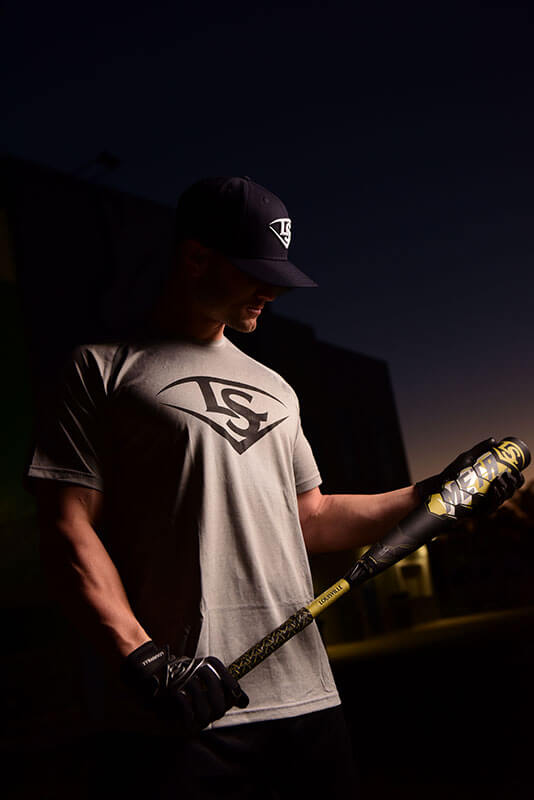 A male baseball player poses with a bat in a darkly lit photograph.
