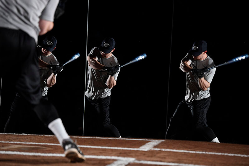 A darkly lit image of a male baseball player swinging a bat in front of mirrors.