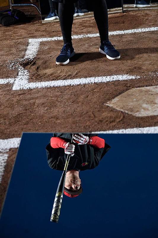 A male baseball player stands in a batter’s box. A mirror on the ground reflects his face and bat.