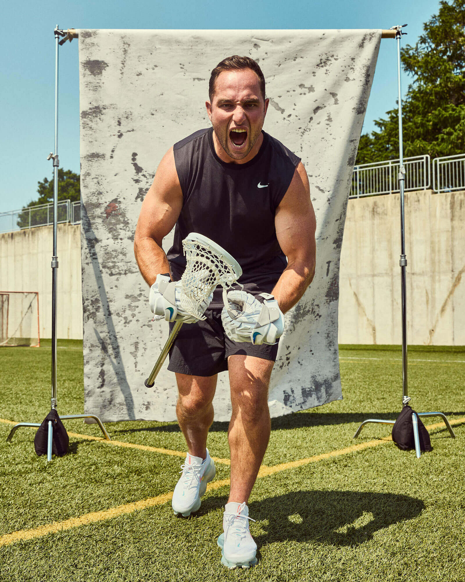 A male lacrosse player strikes a fierce pose on a field in front of a weathered backdrop.
