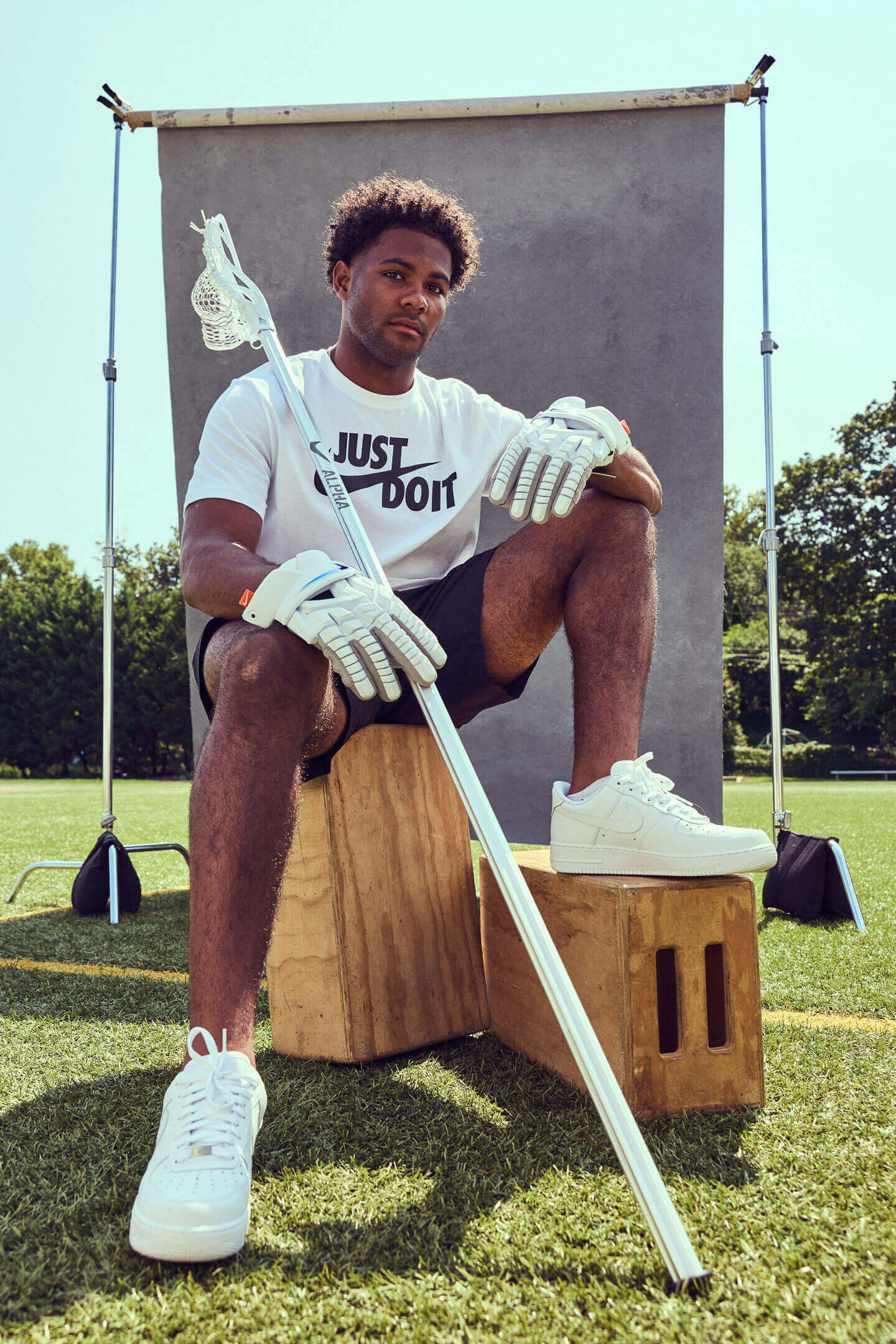 A male lacrosse player stylishly sits on wooden boxes in front of a weathered background on a grass field.