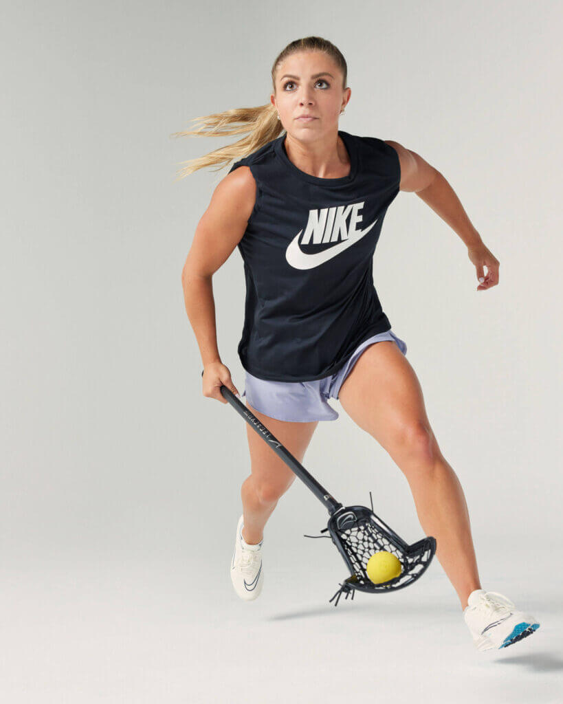 An in-studio action shot of a female lacrosse player running while holding a lacrosse stick.