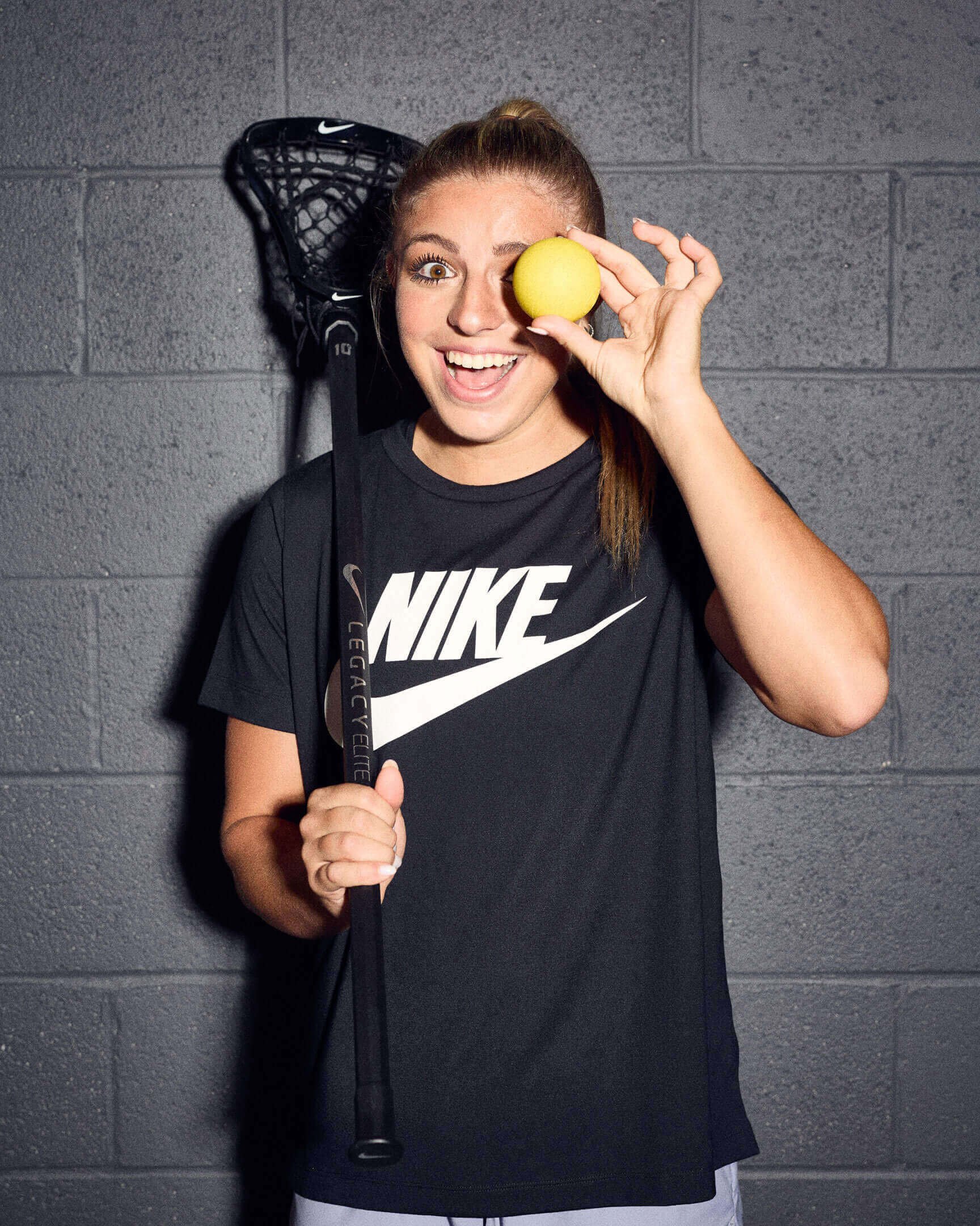 A female lacrosse player poses in a hallway with a lacrosse stick in one hand while holding a bright yellow lacrosse ball in front of one eye with the other hand.