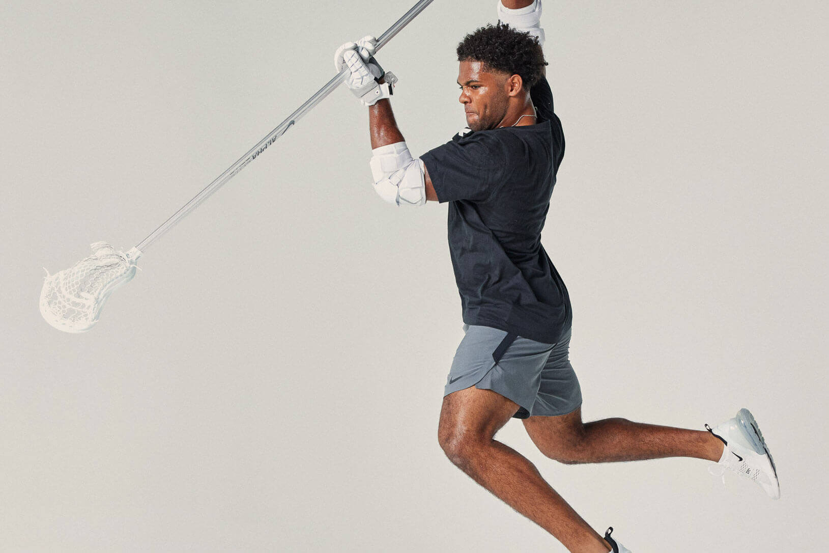 An in-studio action shot of a male lacrosse player leaping with a lacrosse stick in his hands.