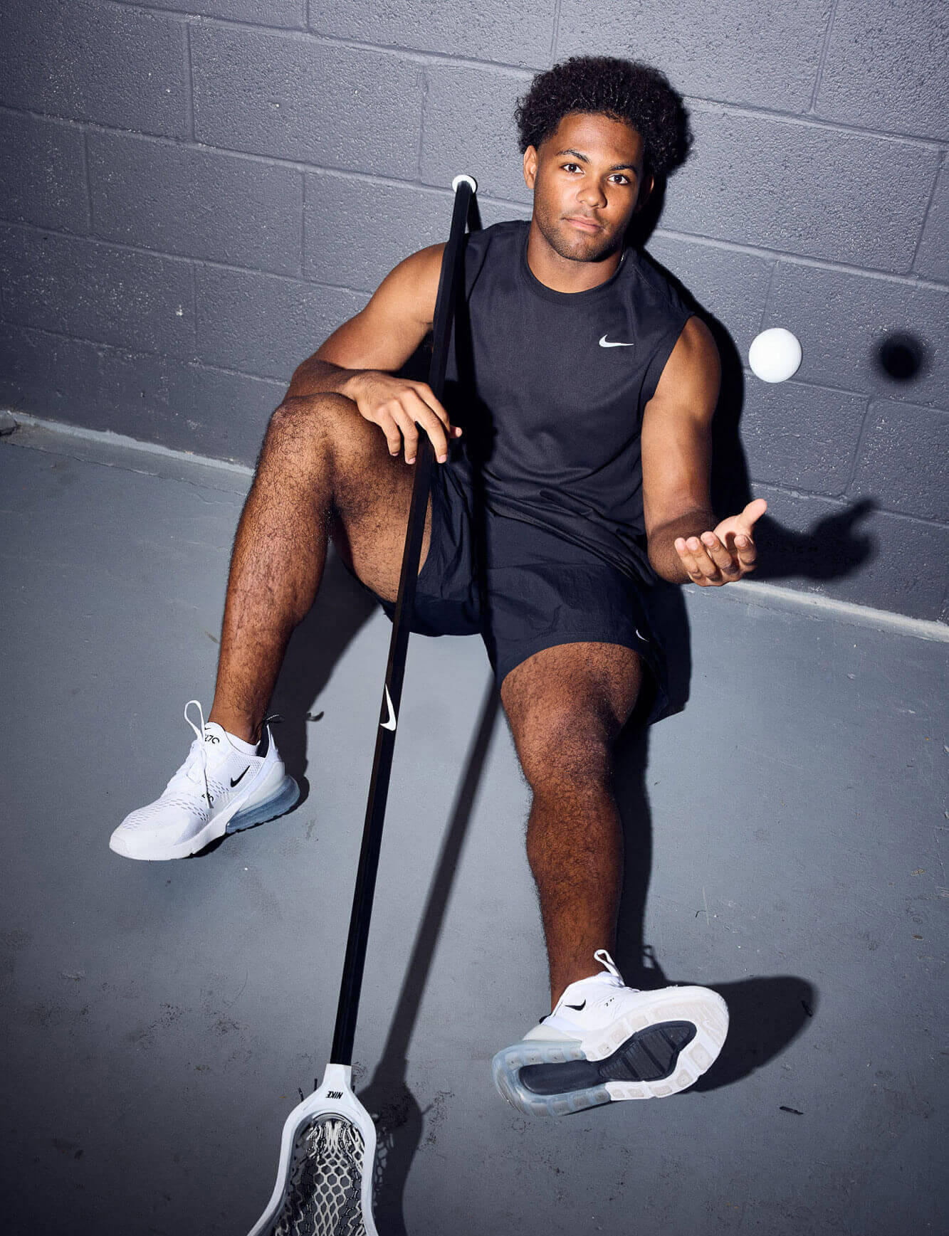 A male lacrosse player sits in a hallway holding a lacrosse stick in one hand and throwing a lacrosse ball up with the other hand.