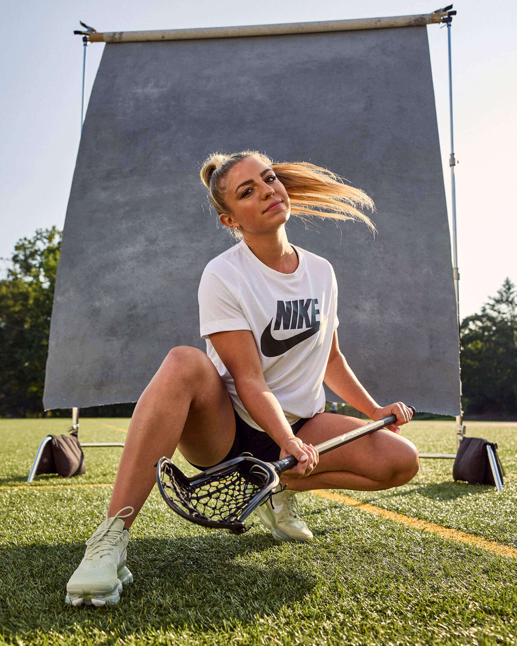 A female lacrosse player stylishly crouches in front of a weathered background on a grass field.