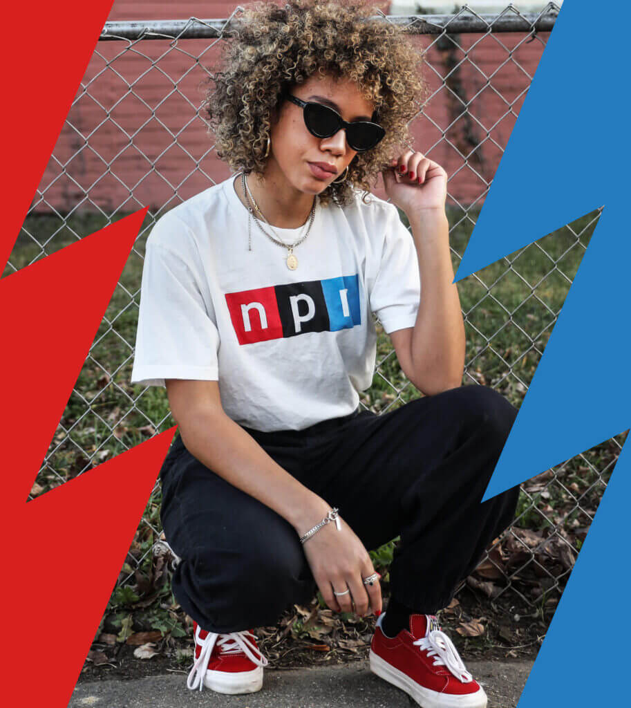 A crouched woman poses outdoors. She’s wearing a white NPR t-shirt, black pants, and sunglasses.
