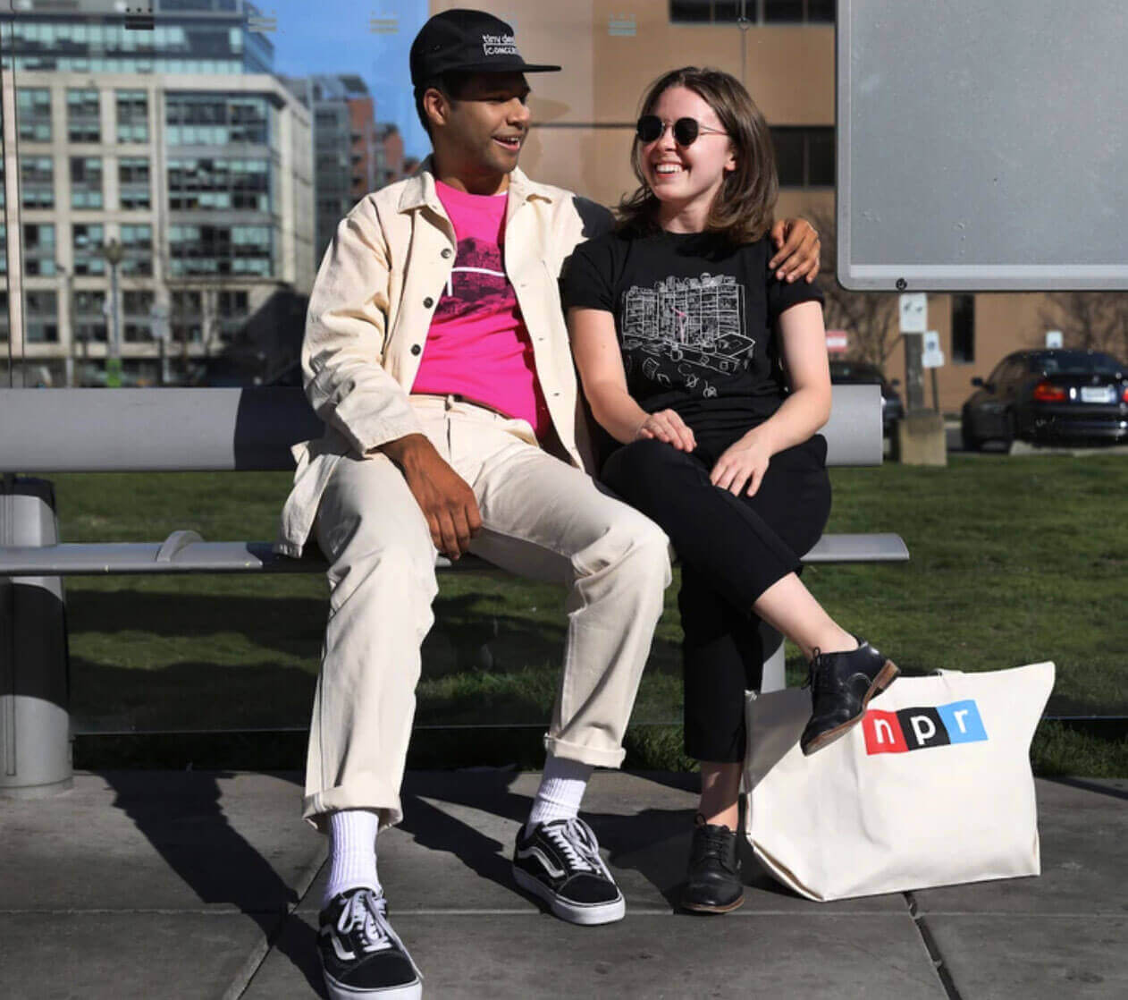 An image of a man and a woman sitting on a park bench outside wearing Tiny Desk t-shirts.