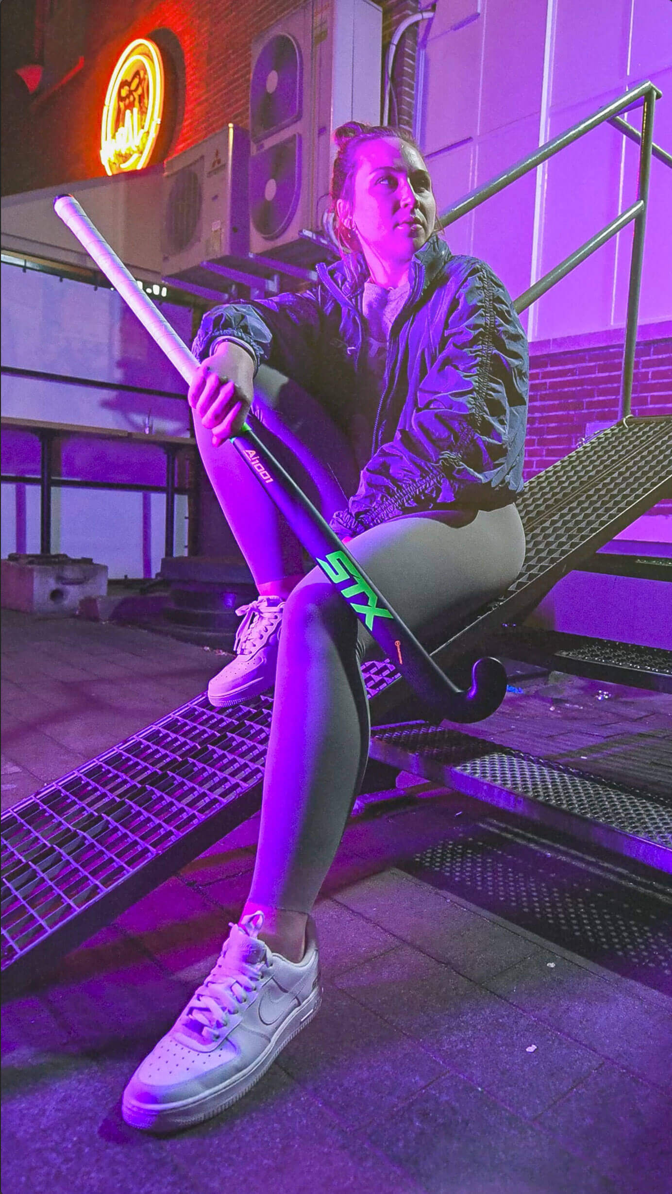 A female field hockey player posing with a field hockey stick sitting on a metal ramp in an urban environment,