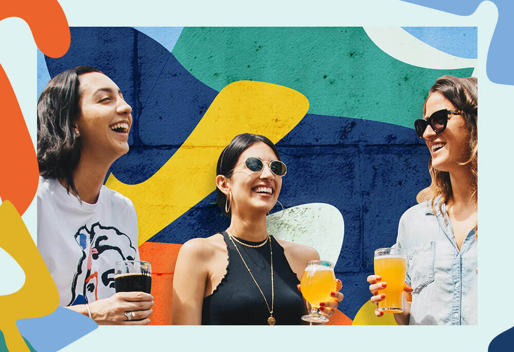 Three women hold pints of beer in front of a colorful mural.