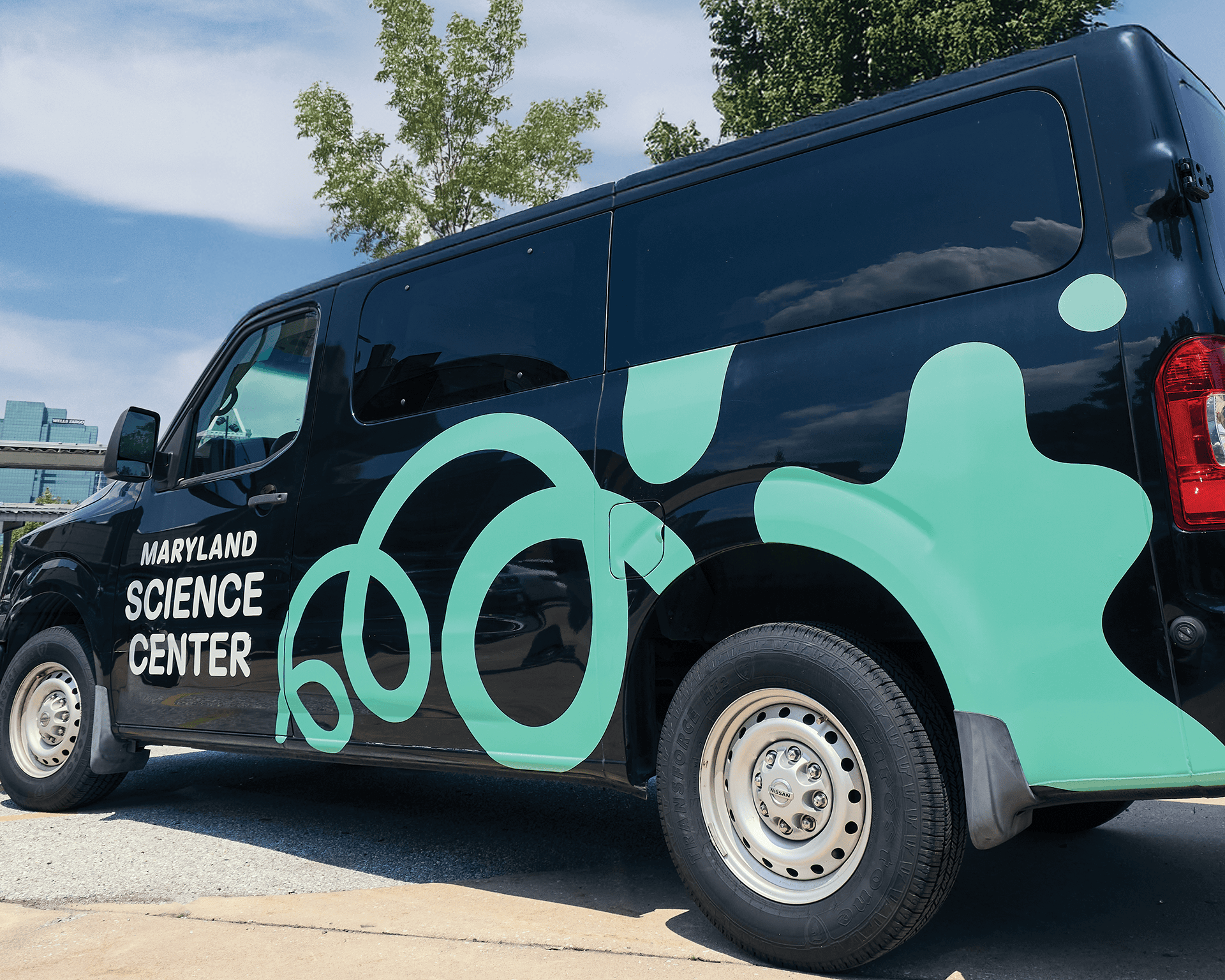 Side profile view of a black transport van parked outside, featuring a large teal Maryland Science Center logo graphic and organic blob shapes wrapped across the side and rear panels.