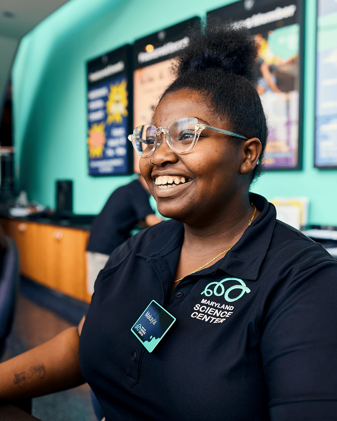 A smiling female staff member named Makayla wearing clear glasses and a black polo shirt embroidered with the teal Maryland Science Center logo.