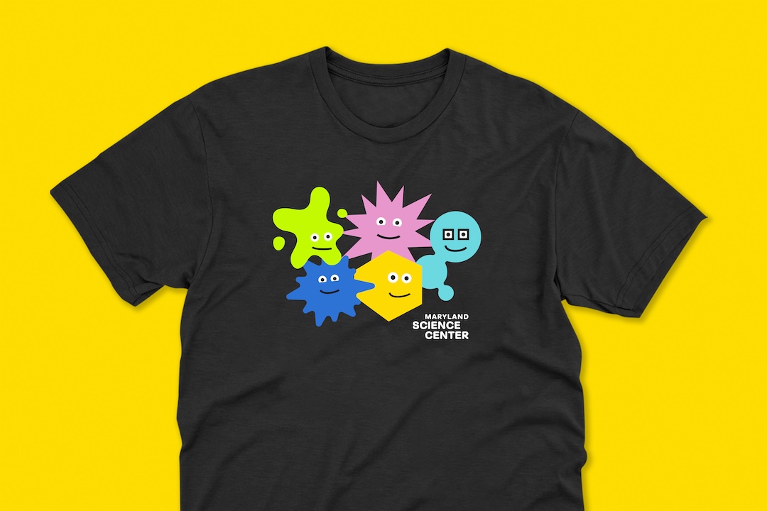 A black t-shirt displayed against a vibrant yellow background, featuring an assortment of colorful, stylized geometric cartoon characters with friendly faces above the white text 