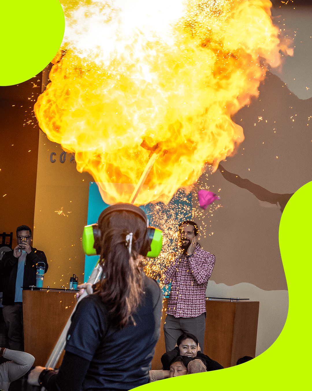 A live science demonstration at the museum where a presenter wearing green noise-canceling headphones uses a tool to create a massive fireball explosion indoors, while audience members watch in amazement. A lime-green organic graphic border frames the image.