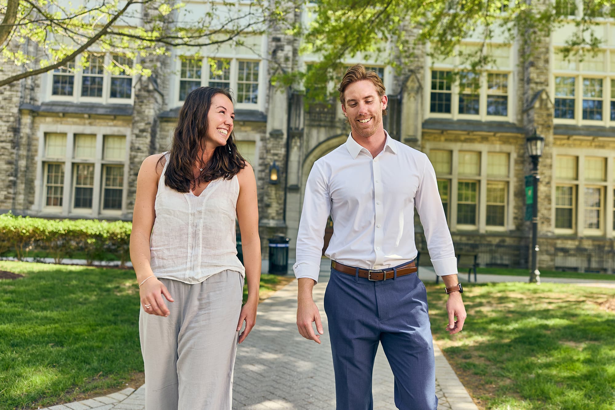 Two students, dressed in business-casual attire, smiling and laughing together as they walk along a paved campus pathway in front of a stone academic building.