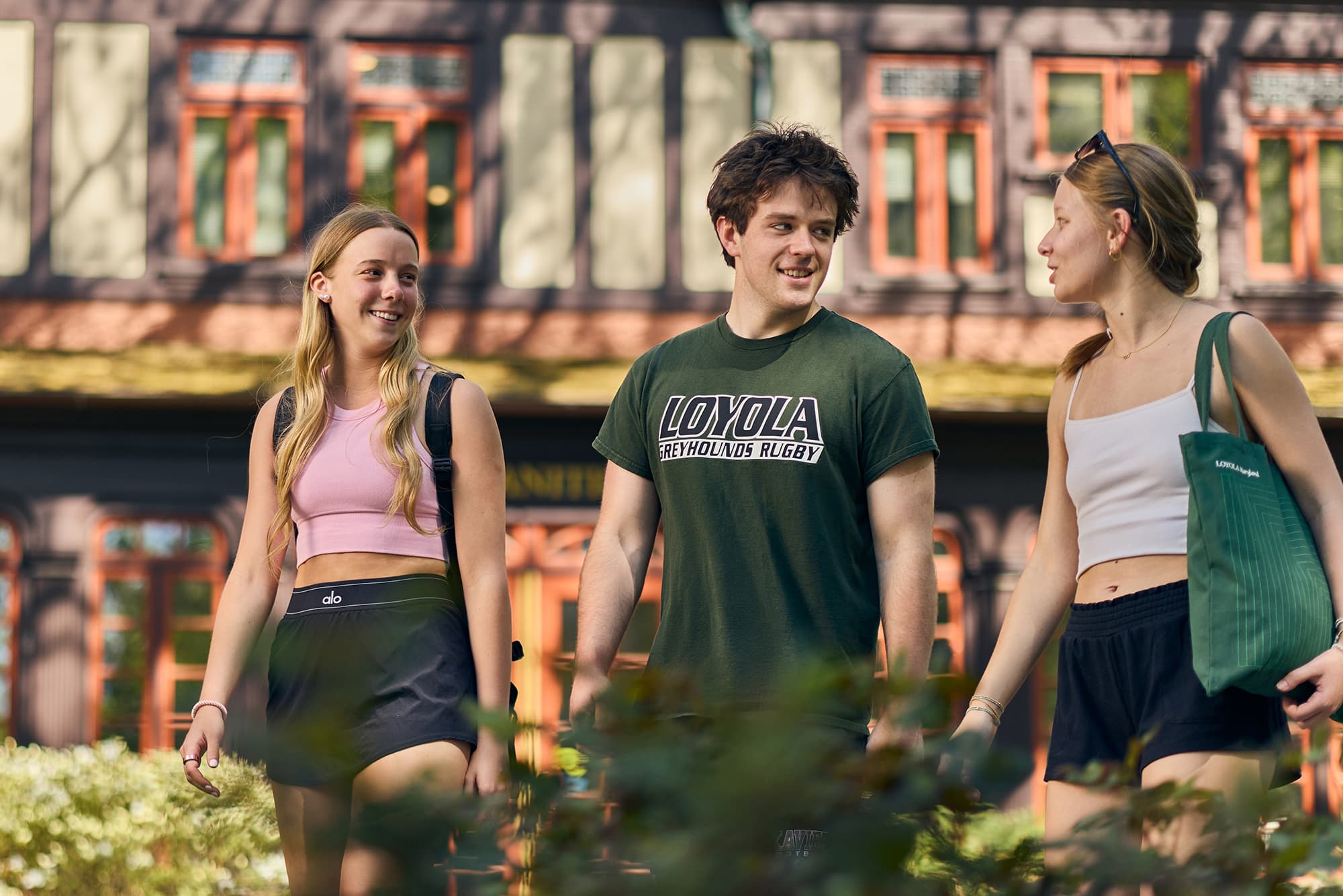 Three students talking and walking together across campus outdoors in front of a Tudor-style building, with the student in the center wearing a green Loyola Greyhounds Rugby t-shirt.