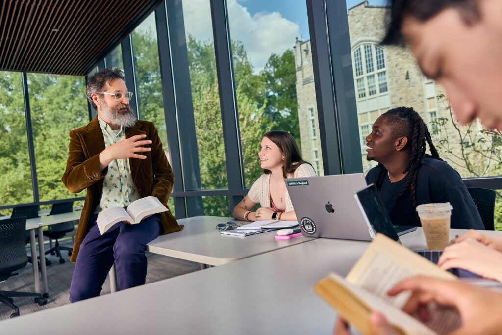 A professor with a beard, wearing a brown jacket and holding an open book, sits on the edge of a table while leading a class discussion with attentive students in a bright room with large glass windows.