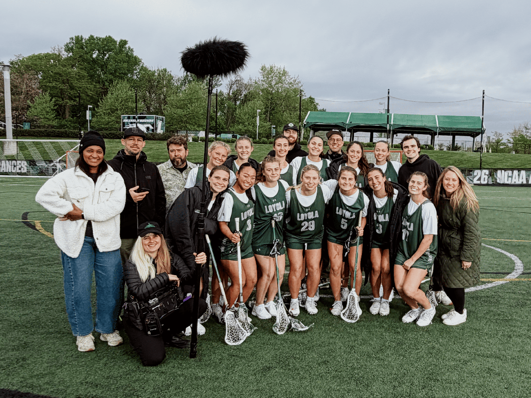 Behind-the-scenes group photo on an athletic turf field showing the Loyola University Maryland women's lacrosse team posing with lacrosse sticks alongside a video production crew holding audio and camera gear.