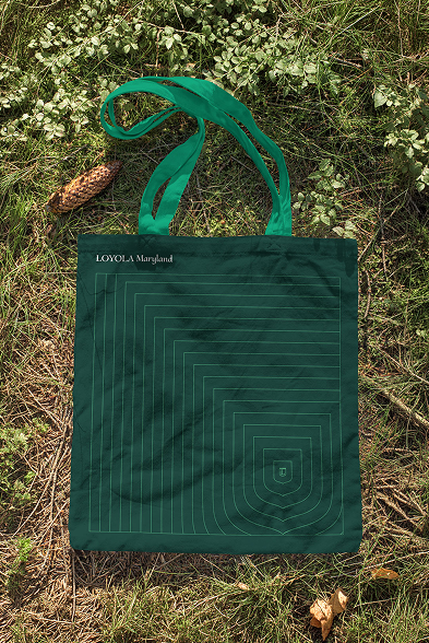 A dark green Loyola Maryland tote bag printed with a geometric shield line graphic laying flat on outdoor grass.