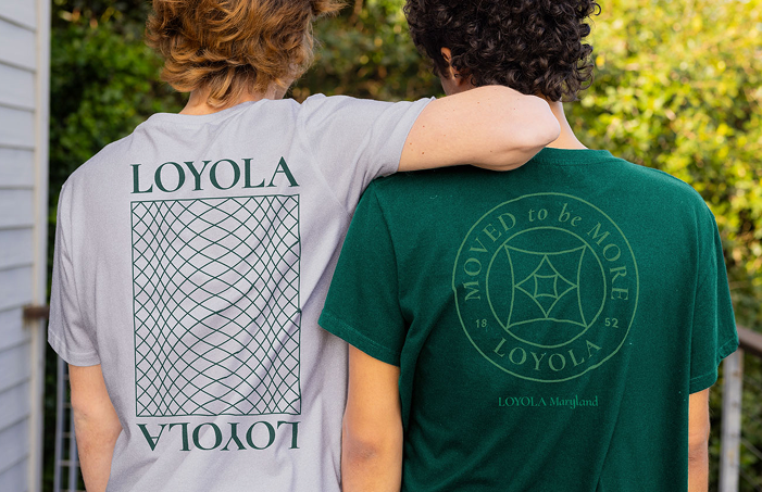 Rear view of two friends with arms around each other's shoulders, wearing grey and dark green t-shirts printed with Loyola graphic designs and the tagline 