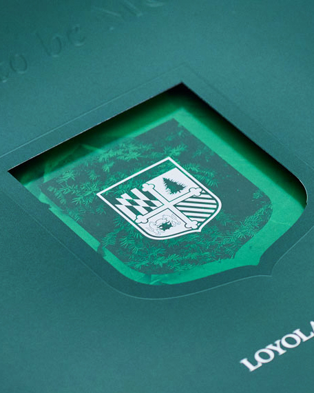 Close-up of the Loyola University Maryland viewbook cover showing a die-cut shield opening that reveals a vibrant green university crest printed with pine needles and a cross emblem.