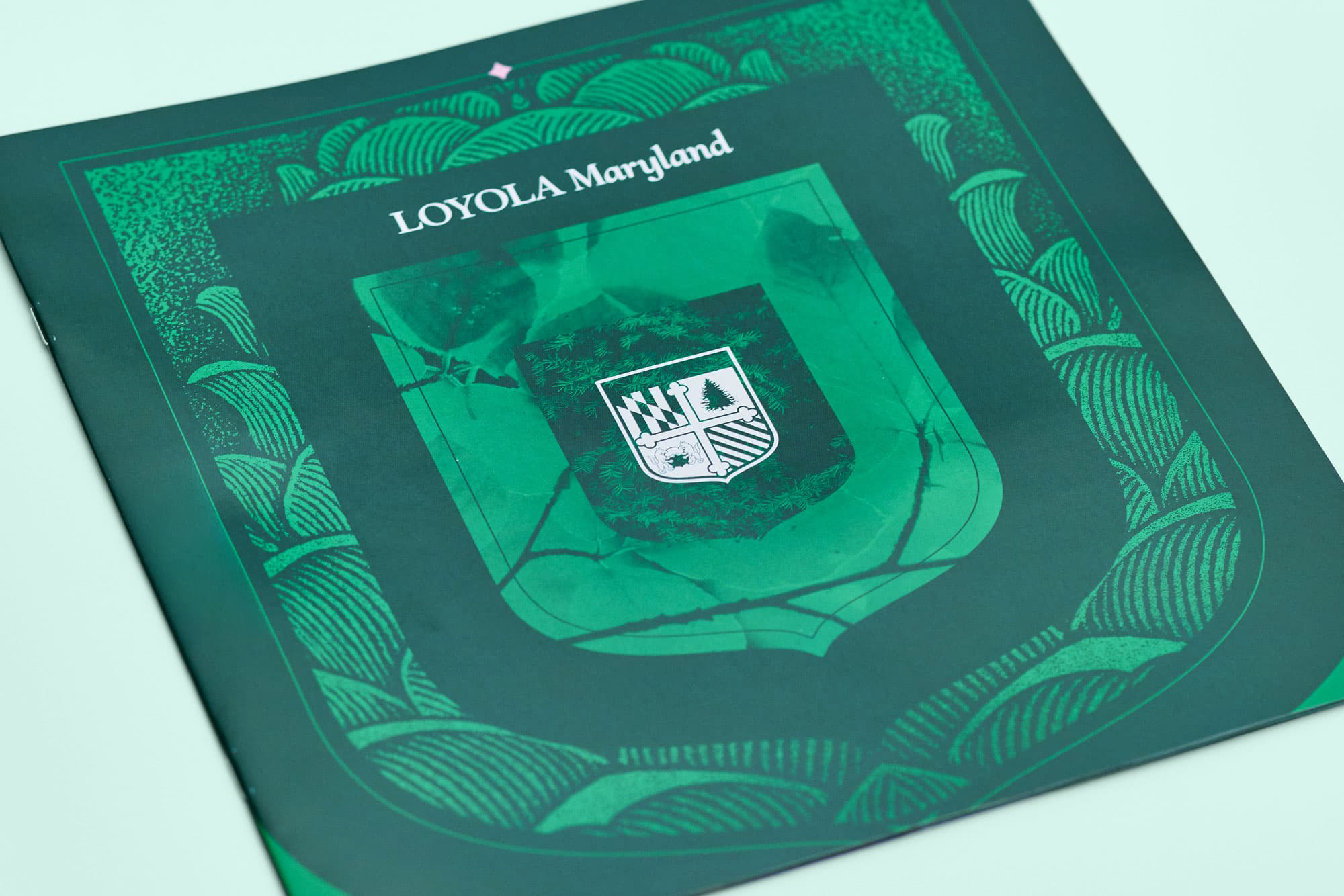 Front cover of a green Loyola Maryland booklet displaying an intricate botanical frame illustration around the center shield logo and university name.