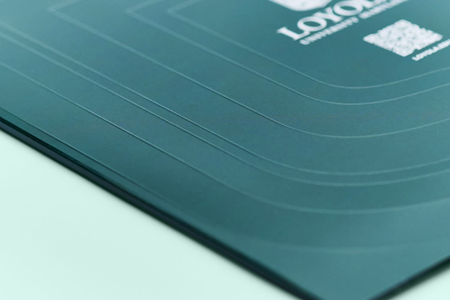 Close-up detail of debossed arch lines along the corner edge of a dark green Loyola University Maryland viewbook folder.