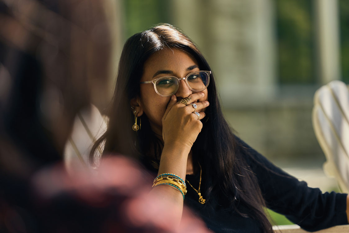 Close-up shot of a smiling student wearing clear-framed glasses, gold jewelry, and stacked bracelets, laughing gently with her hand over her mouth during an outdoor conversation.