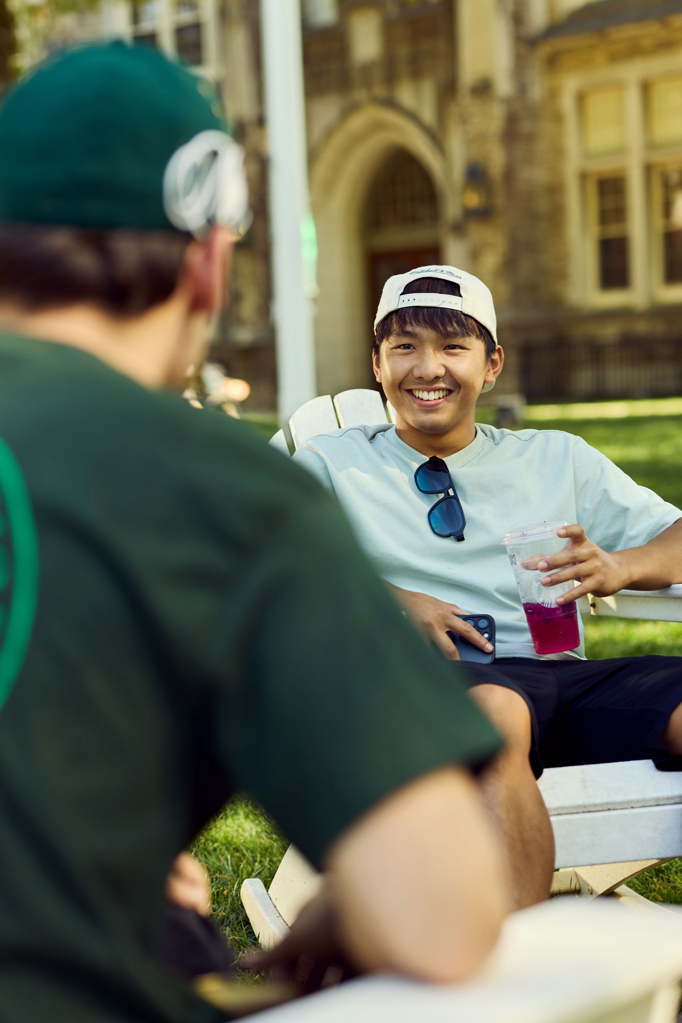 A student wearing a white backward baseball cap and sunglasses resting on his shirt collar, holding a drink and smiling while sitting on an outdoor chair across from a friend.