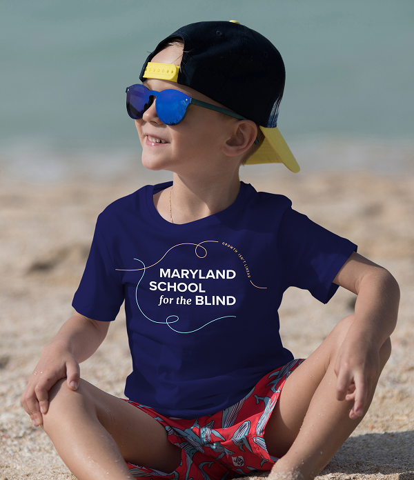 A young boy sitting on a sandy beach wearing sunglasses, a backwards cap, and a navy blue t-shirt featuring the Maryland School for the Blind logo surrounded by curved line art.